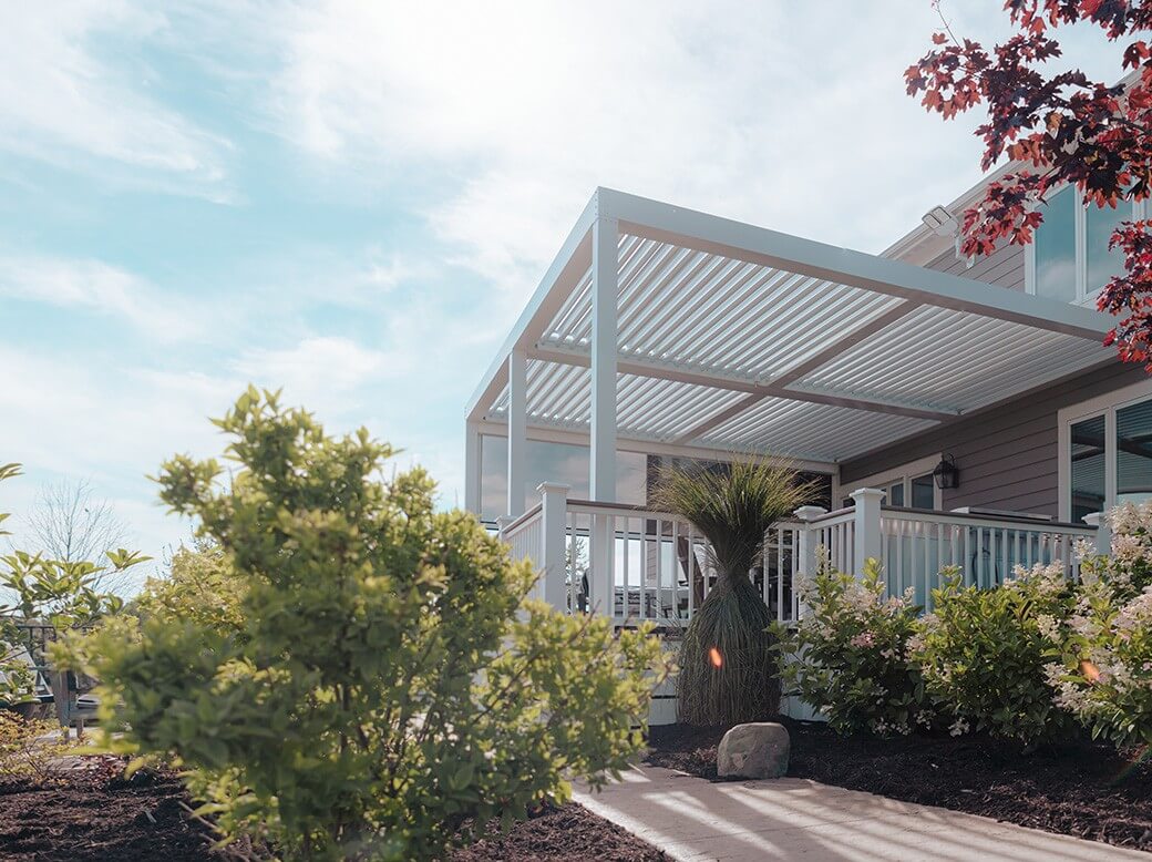 Modern backyard pergola attached to a residential house, featuring a stylish metal frame over a deck enhanced by lush greenery and decorative bushes on a sunny day. The serene setting showcases clear skies and well-maintained landscaping, providing an ideal outdoor relaxation spot.