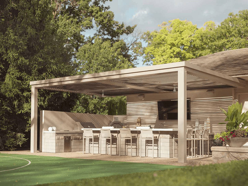 A luxurious outdoor kitchen and dining area under a modern pergola, complete with a fully equipped bar, high stools, and an integrated grill, set against a backdrop of lush greenery and a well-maintained lawn.