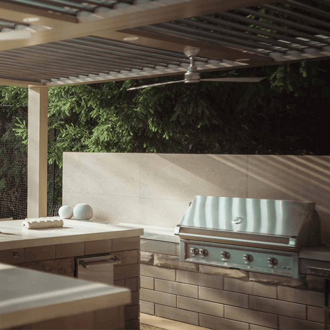 A stylish outdoor kitchen setup featuring a large stainless steel grill built into a stone countertop, with a modern ceiling fan above and lush greenery in the background. The scene captures the essence of luxurious outdoor living and entertaining.