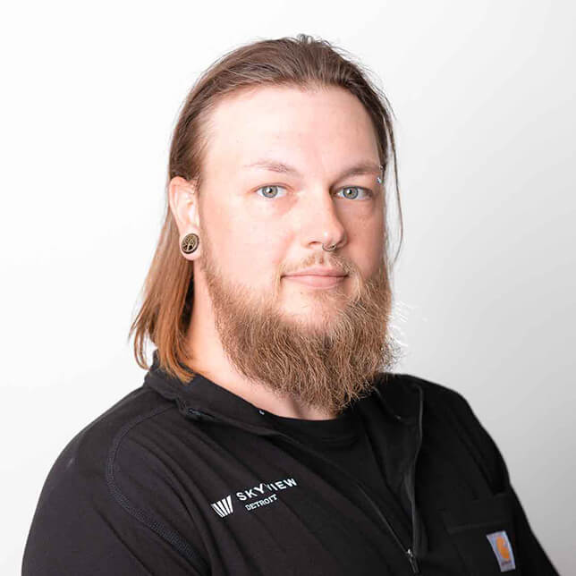 Portrait of a confident man with long hair and beard, wearing a black work shirt labeled "Skyview" and a small earring, set against a plain white background.