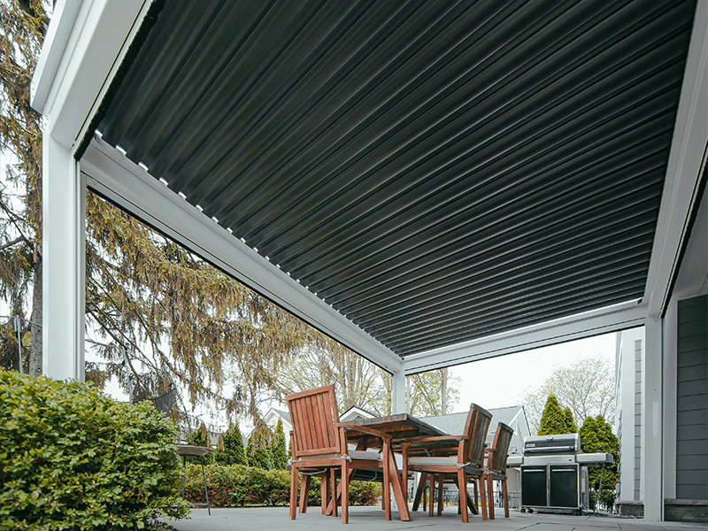 Well-designed outdoor patio area featuring a retractable aluminum roof over a wooden dining set, surrounded by lush greenery and a residential backdrop, creating a comfortable and stylish space for dining and relaxation outdoors.