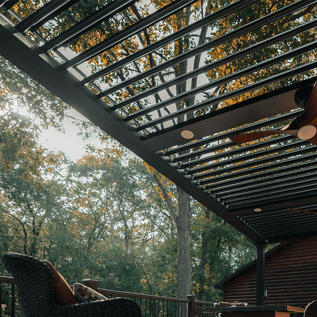 Luxurious outdoor living area featuring a modern pergola with adjustable louvers over comfortable wicker furniture, with a serene view of dense trees in autumn colors.