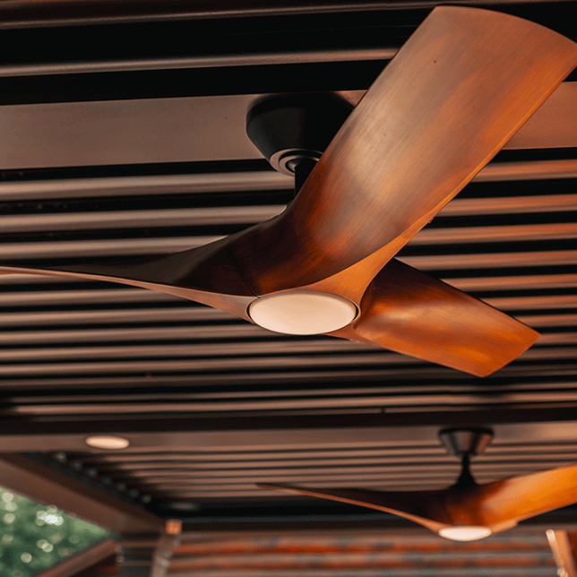 An elegantly designed ceiling fan made of polished wood with three blades, illuminated by warm lighting, is installed against a structured, dark brown ceiling, creating a cozy and modern atmosphere.
