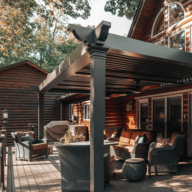 Cozy outdoor sitting area on a wooden deck featuring a modern pergola, comfortable wicker furniture, and a picturesque log cabin in the background, surrounded by trees. This setting exemplifies a serene outdoor living space suitable for relaxation and entertaining.