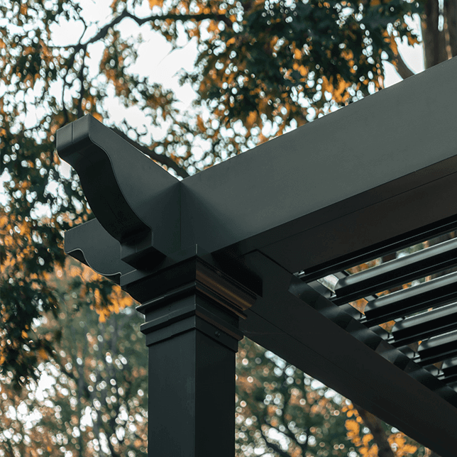 Close-up view of a modern outdoor pergola with dark metallic beams and a stylish gutter system set against blurred autumn foliage in the background.