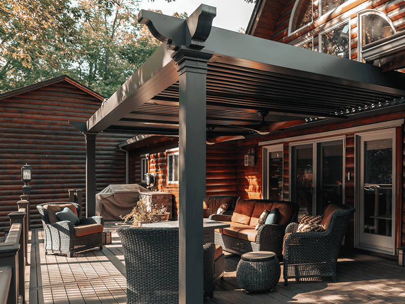 Cozy outdoor living space on a wooden deck featuring comfortable rattan furniture and a stylish pergola, set against the warm, natural wood exterior of a log cabin surrounded by trees.