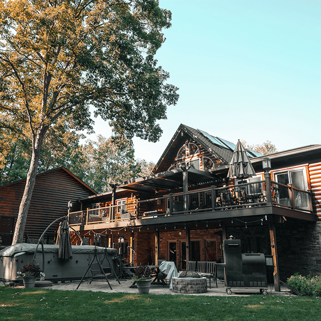 A rustic log cabin home surrounded by lush trees, featuring a spacious outdoor deck with multiple levels and leisure amenities including a hot tub and a large grill, set against a clear blue sky.