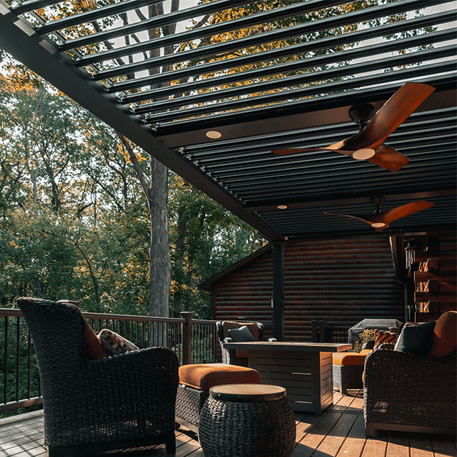 Elegant outdoor seating area on a wooden deck featuring a modern pergola with louvers, surrounded by lush trees, enhanced with cozy furniture and a ceiling fan for comfort. This tranquil setting offers a perfect spot for relaxation in a natural environment.