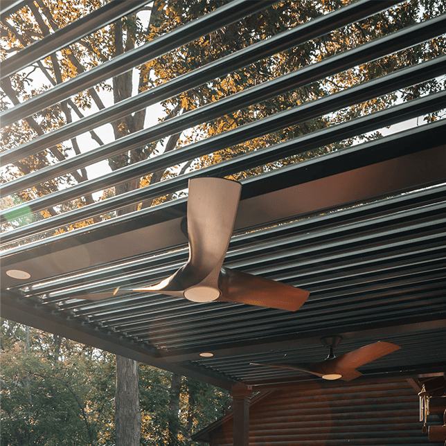 Outdoor patio area with modern ceiling fans integrated into a wooden slatted roof structure, filtering sunlight through the surrounding trees to create a serene ambiance.