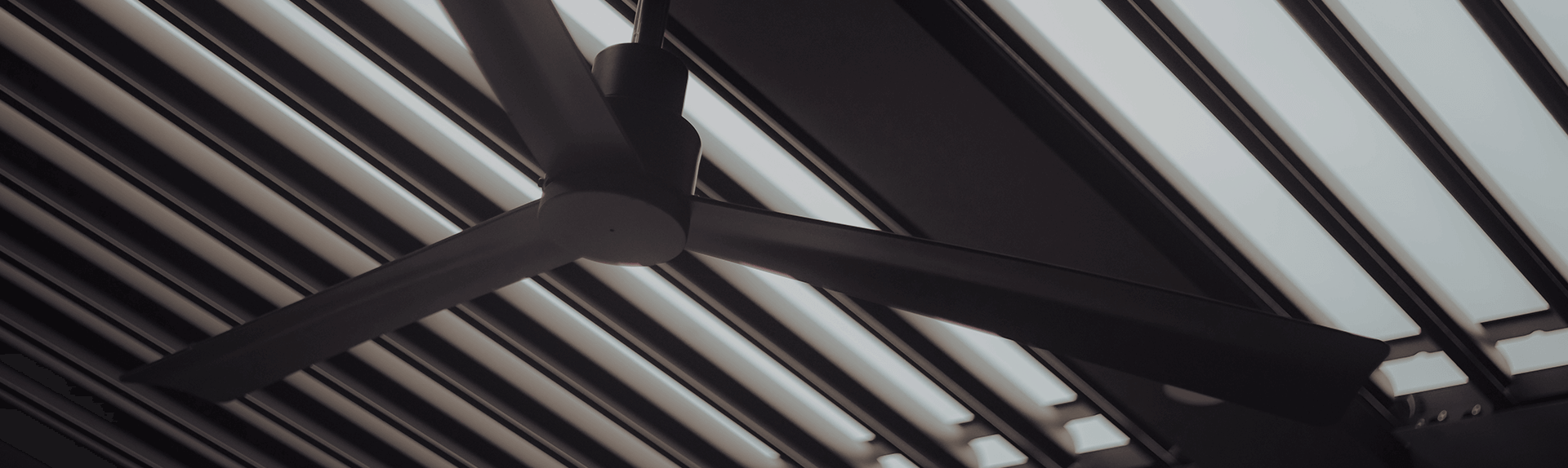 A modern ceiling fan with three sleek blades suspended under a linear patterned roof structure in a moody, monochrome setting. The image highlights the minimalist design and architectural aesthetics common in contemporary interior spaces.
