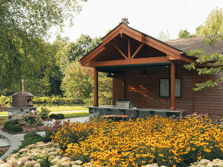 A cozy wooden pavilion with a large bronze pizza oven and outdoor seating area surrounded by lush gardens full of blooming yellow and white flowers under a clear sky.
