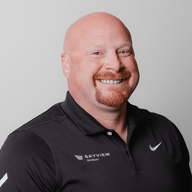 A bald man with a friendly smile wearing a black Nike polo shirt with a logo reading "SKYVIEW Detroit" on the left chest directs a warm gaze towards the camera. His professional appearance conveys approachability and confidence.
