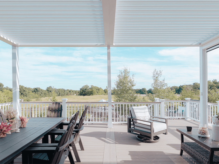 View from a spacious and neatly organized backyard covered patio with stylish outdoor furniture overlooking a scenic, tree-lined landscape under a clear sky.
