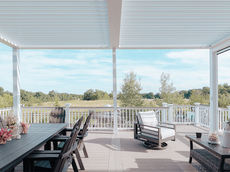 View from a spacious and neatly organized backyard covered patio with stylish outdoor furniture overlooking a scenic, tree-lined landscape under a clear sky.