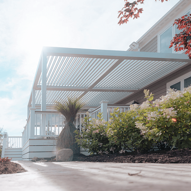 Modern outdoor patio area featuring a stylish open louvered roof system providing adjustable sunlight, equipped with cozy seating arrangements including recliners and a sofa, offering a serene view of a clear sky and lush greenery in a tranquil setting.