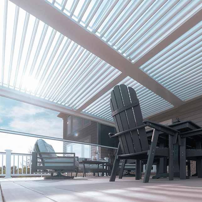A serene view of a residential home's outdoor pergola and decking area, showcasing a stylish metal structure shaded by slatted beams with surrounding lush plants under a clear sky.