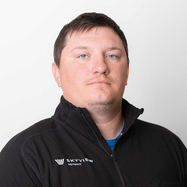 Portrait of a man wearing a black Skyview Detroit jacket over a blue shirt, staring directly at the camera with a slight expression, set against a plain light background.