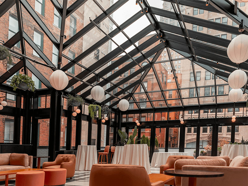 A cozy sunlit nook features a large, plush brown sofa adorned with various cushions, flanked by large windows showcasing a garden view, creating an inviting and warm space for relaxation.