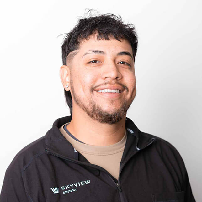 A cheerful young man with a light beard smiles in a professional portrait, dressed in a dark jacket labeled "Skyview Detroit." His friendly demeanor and casual hairstyle reflect a positive work environment.