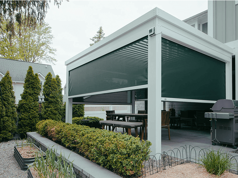 Modern rooftop patio featuring a wooden deck with a stylish dining set and a comfortable lounge area, surrounded by clear glass balustrades under a wooden pergola, offering a panoramic view of a serene suburban landscape on a sunny day.