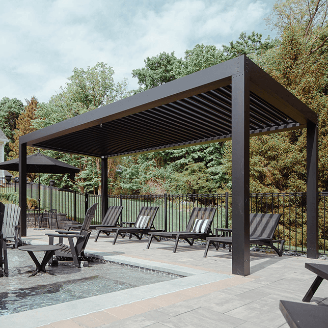 A modern outdoor lounge area featuring a sleek black pergola over multiple sun loungers, adjacent to a swimming pool and fenced yard, set against a backdrop of a residential building and lush greenery.