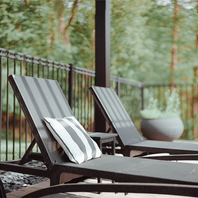 A modern outdoor patio area featuring a sleek dark-colored pergola over lounge chairs, with a well-kept grass lawn and residential buildings in the background under a clear sky. The design integrates contemporary style with comfort, suited for rest and gatherings.