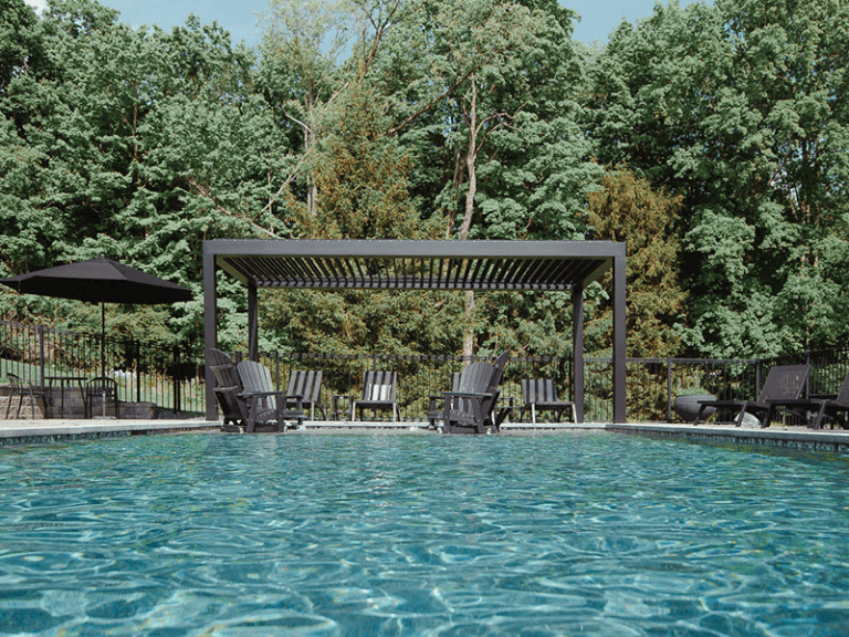 A serene outdoor pool area with crystal-clear blue water surrounded by dark patio furniture under a modern pergola, set against a backdrop of lush green trees.
