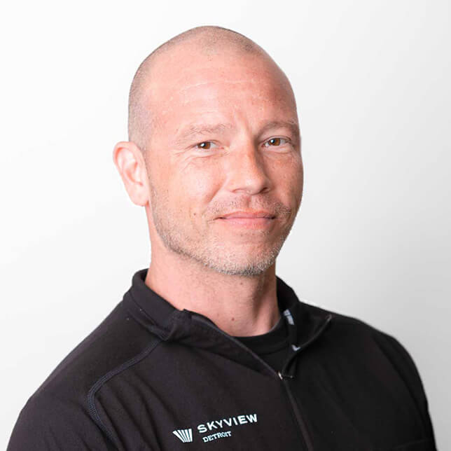 A portrait of a middle-aged Caucasian man with a shaved head and subtle facial hair, wearing a black zippered jacket with the logo "Skyview Detroit" embroidered on the left side; he smiles lightly with a white background.