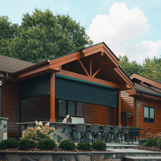 A modern outdoor seating area with a large motorized retractable screen, surrounded by a stone bar countertop and wooden high chairs, set against the backdrop of a log cabin with exposed wooden beams and lush greenery.