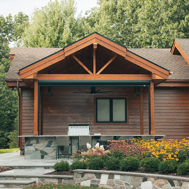 A modern outdoor kitchen under a wooden ceiling structure featuring black support beams, built-in lighting and ceiling fans, with a view of lush green trees and a clear sky.