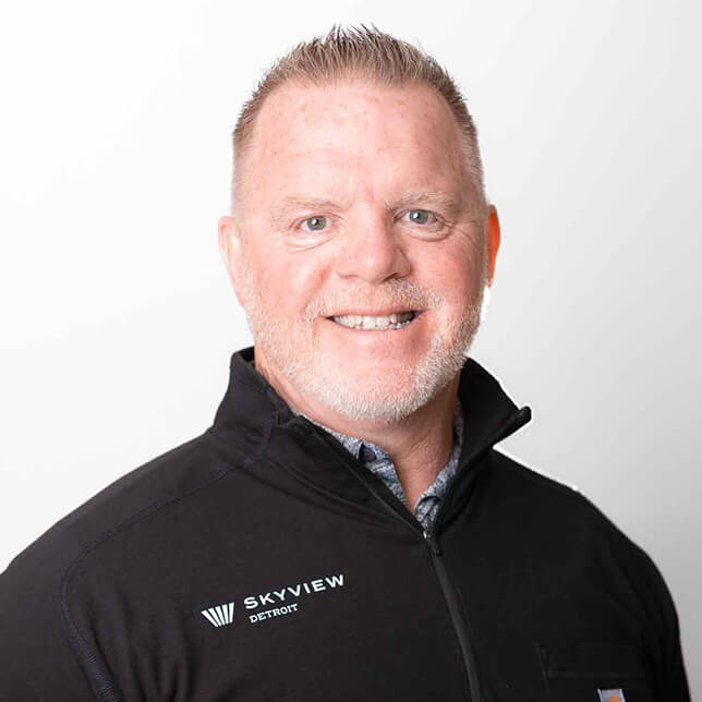 A professional portrait of a smiling man with short, cropped gray hair wearing a black jacket embroidered with the logo "Skyview Detroit". He stands against a plain white background, giving a friendly and approachable appearance.