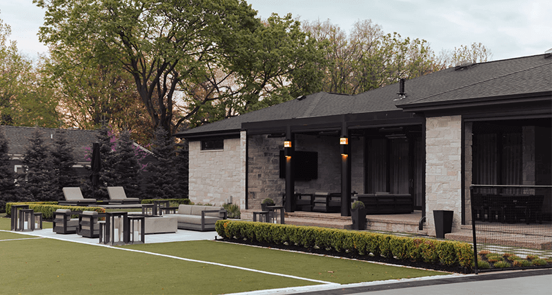 A luxurious outdoor patio featuring a long black dining table under a pergola with adjustable roof slats, complemented by an outdoor kitchen to the left; in the background, a meticulously landscaped yard with a soccer goal and basketball court.