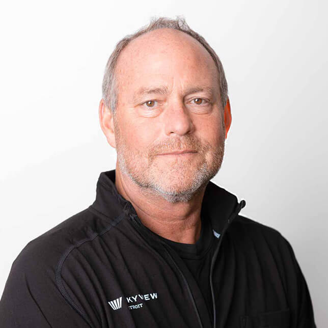 Middle-aged man with a slight smile, wearing a black zippered jacket with a logo on the left chest, standing against a plain white background. His appearance is casual-professional, emphasizing an approachable and experienced demeanor.