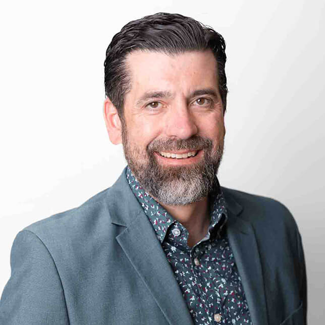 A professional headshot of a smiling man with a neatly trimmed beard, dressed in a grey blazer over a patterned shirt, set against a light gray background. His pleasant expression and warm demeanor give a welcoming impression.