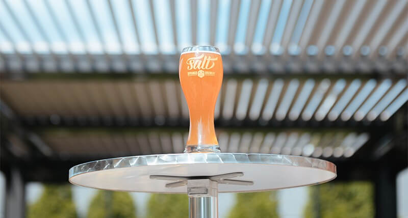 A glass of amber-colored beer branded with "Salt" on a glass outdoor table, with a blurred architectural canopy and clear sky in the background, conveying a refreshing outdoor setting at a brewery.