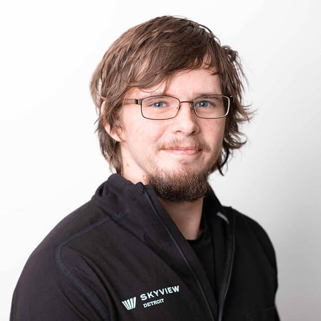 A man with tousled brown hair and rectangular glasses smiles gently, wearing a black jacket with a SkyView Detroit logo. He stands against a clean white background that highlights his approachable demeanor.