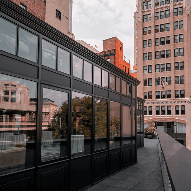 A contemporary rooftop dining area enclosed by a glass and black metal structure, illuminated by soft interior lighting and surrounded by urban buildings, with a single tree in a large pot adding a touch of nature.