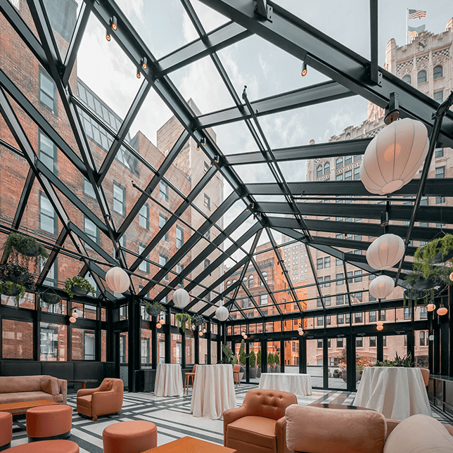 Elegant lounge area under a modern glass roof with dark metal beams, featuring stylish terracotta chairs and sofas arranged around a central bar lit by warm lights, set against a dark brick wall with marble accents.