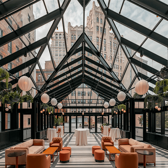 Elegant modern event space with a glass ceiling and black frames, furnished with stylish orange sofas and wooden coffee tables, and adorned with white paper lanterns and greenery, offering a view of the surrounding urban landscape.