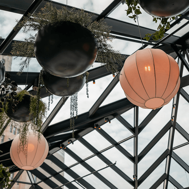 View looking up at a modern glass roof structure with black metal framing, equipped with small orange lights, set against a backdrop of a traditional brick building. The contrasting architectural styles highlight the blend of new and old design elements.