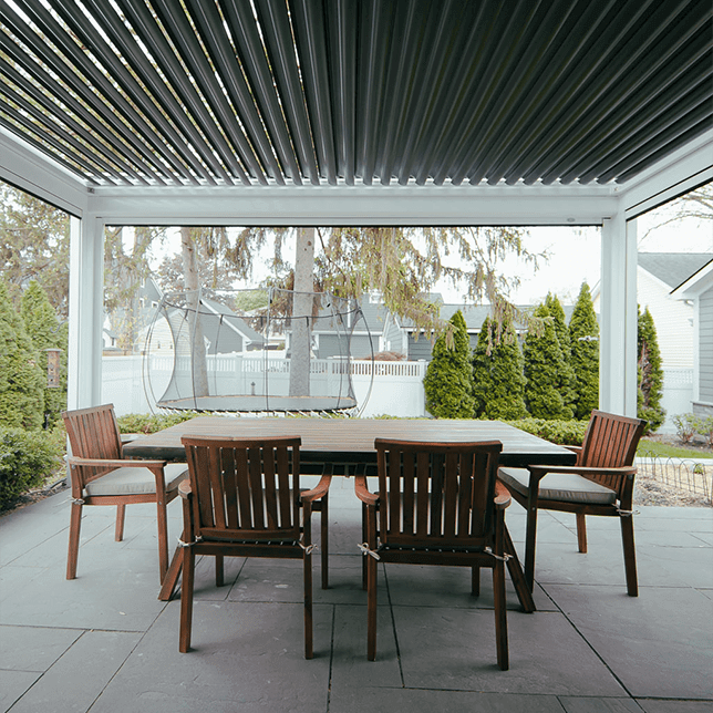 Four wooden chairs gathered around a table under a pergola with a louvered roof, set in a serene garden with tall trees visible in the backdrop, evoking a peaceful outdoor dining or relaxation area.