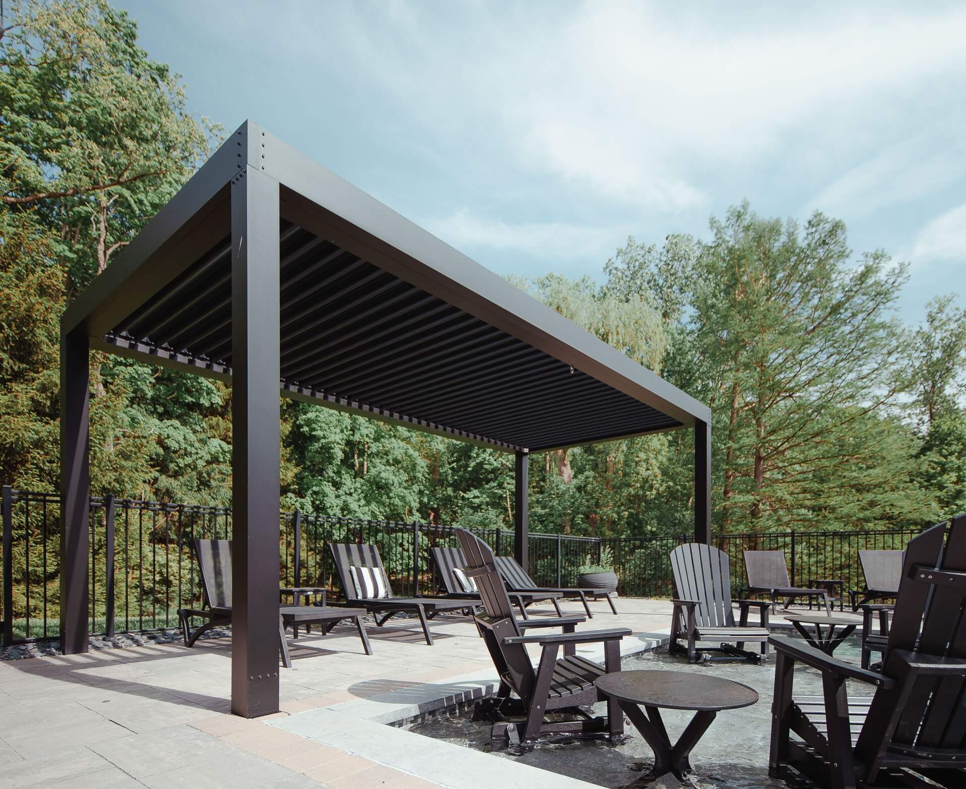 Modern outdoor relaxation area featuring a large, dark-colored pergola with adjustable louvers, surrounded by multiple lounge chairs and small tables, set on a textured patio overlooking a lush green backdrop.