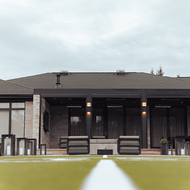 A luxurious outdoor living space featuring a modern patio covered by a dark pergola with integrated lighting, complete with stylish lounge furniture and a stone wall background, set in a well-manicured garden.