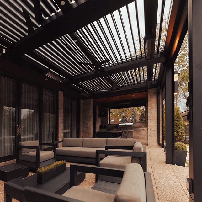 Modern outdoor patio area featuring comfortable seating under a sleek, adjustable louvered roof with integrated heating elements, set against a backdrop of lush trees in a tranquil suburban setting.
