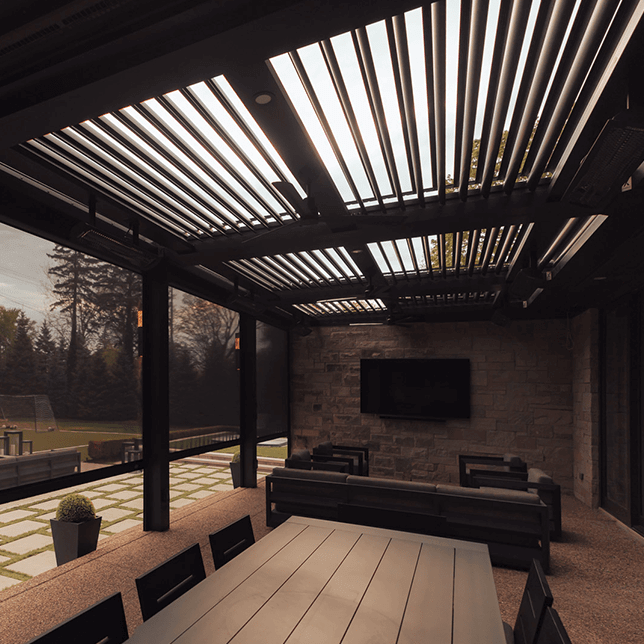 Modern outdoor living space featuring an elegantly mounted flat-screen TV on a textured stone wall, surrounded by sleek black furniture under a stylish pergola with open louvers, overlooking a manicured lawn.