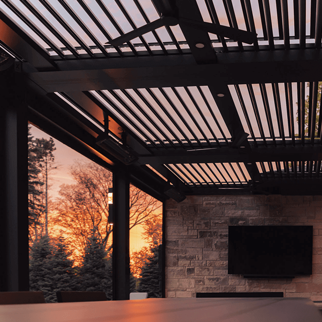 Luxurious outdoor living space featuring a modern pergola with adjustable slats, elegant stone wall with a mounted television, and contemporary furniture, set against a backdrop of lush greenery visible through floor-to-ceiling glass walls.