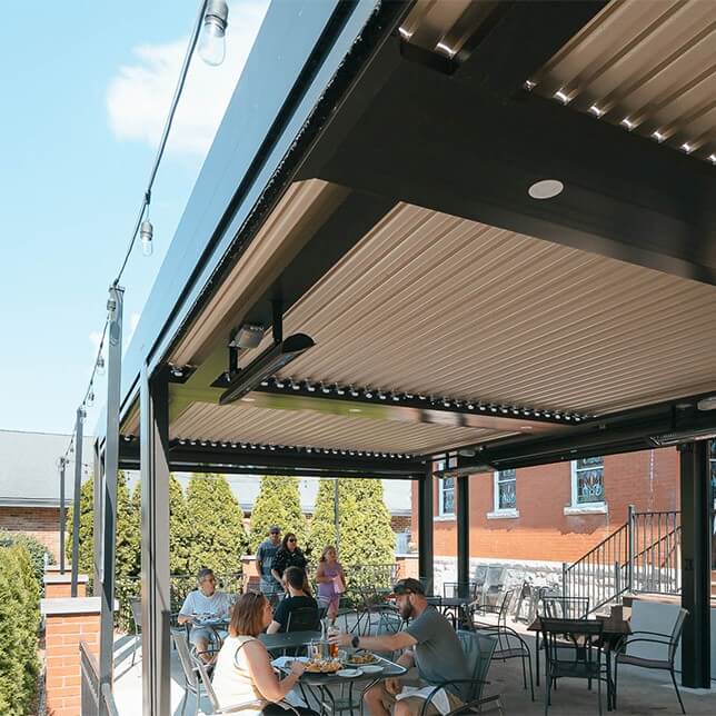 Outdoor dining area under a retractable awning at a modern restaurant, with people enjoying meals and conversations on a sunny day. The setting features stylish outdoor furniture and string lights above, contributing to a comfortable and inviting ambience.