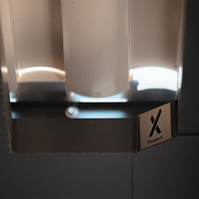 Close-up view of a modern Pergola X structure featuring cylindrical components and a metal label clearly displaying the brand name. The lighting creates warm highlights on the gray and white tones of the architecture.