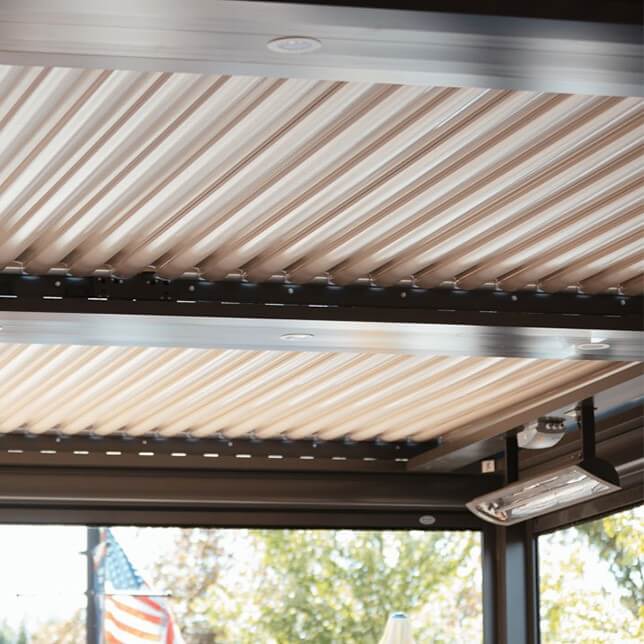 Modern retractable awning system installed on a deck, featuring beige fabric stretched between metallic tracks under a clear blue sky, with visible lighting fixtures and a distant view of an American flag.
