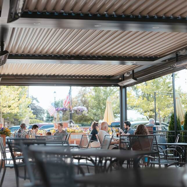 Outdoor patio dining at a restaurant with people casually seated at tables, enjoying meals under a retractable awning with floral arrangements and a view of an American flag outside.